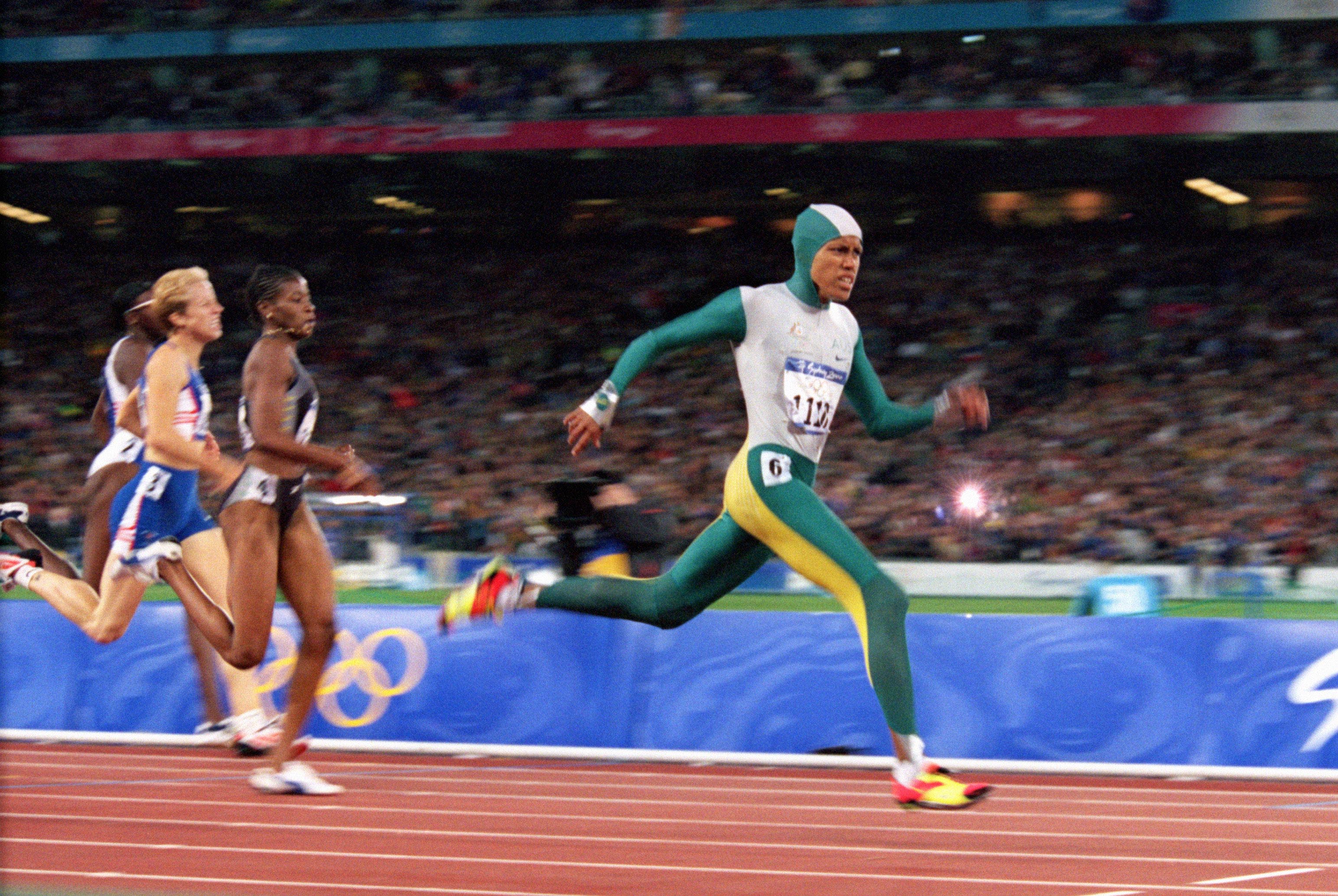 Cathy Freeman in action at the 2000 Sydney Olympics,
                wearing her iconic green and gold body suit and white cap,
                dominating the track event with fierce competition in the
                backdrop.